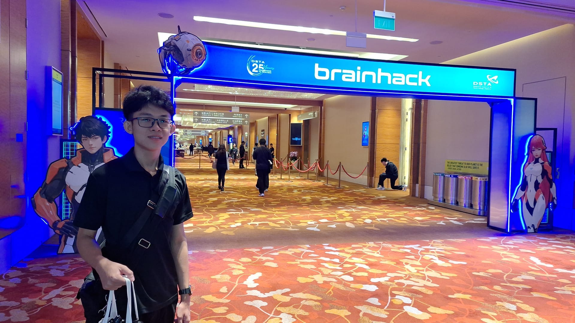 Person in glasses standing at the blue-lit DSTA BrainHack 2025 entrance between science-fiction character displays.