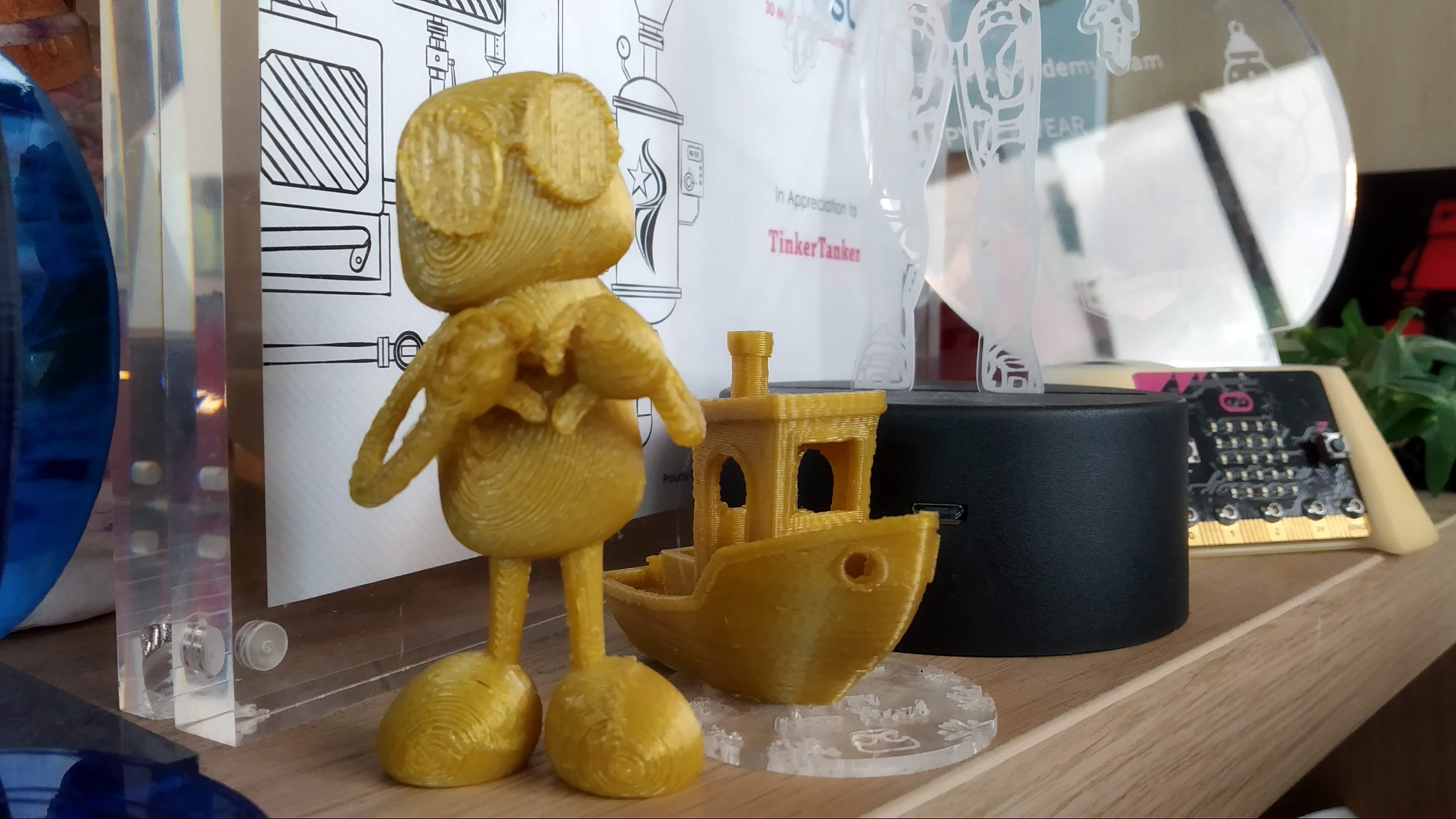 The golden 3DBenchy and TKRobot, together on top of the company KALLAX shelves. (Fun fact: The acrylic piece underneath the 3DBenchy is a draft of the Tinkerfest bauble)