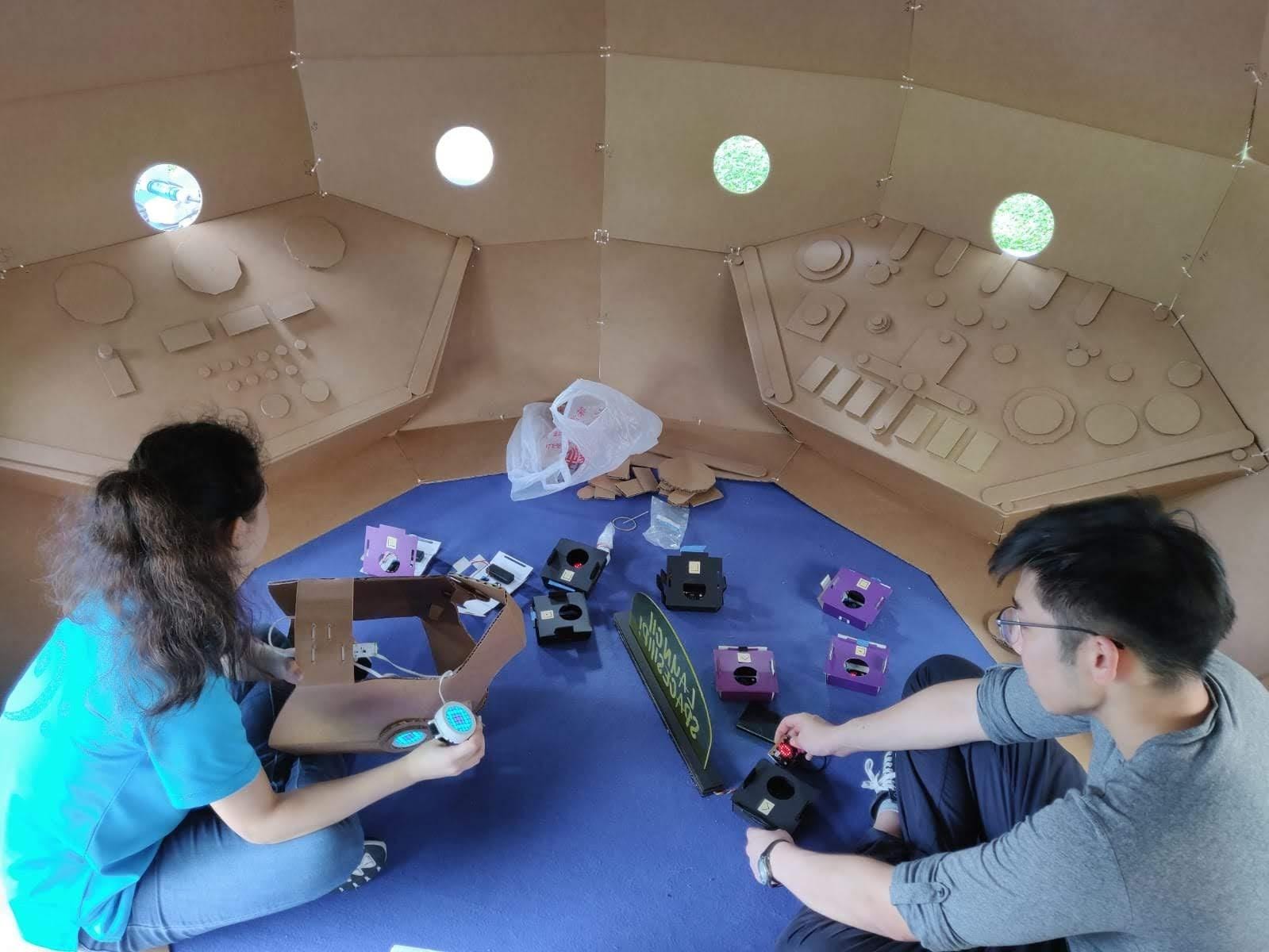 Two people assemble cardboard target boxes with taped flaps around the light sensors.