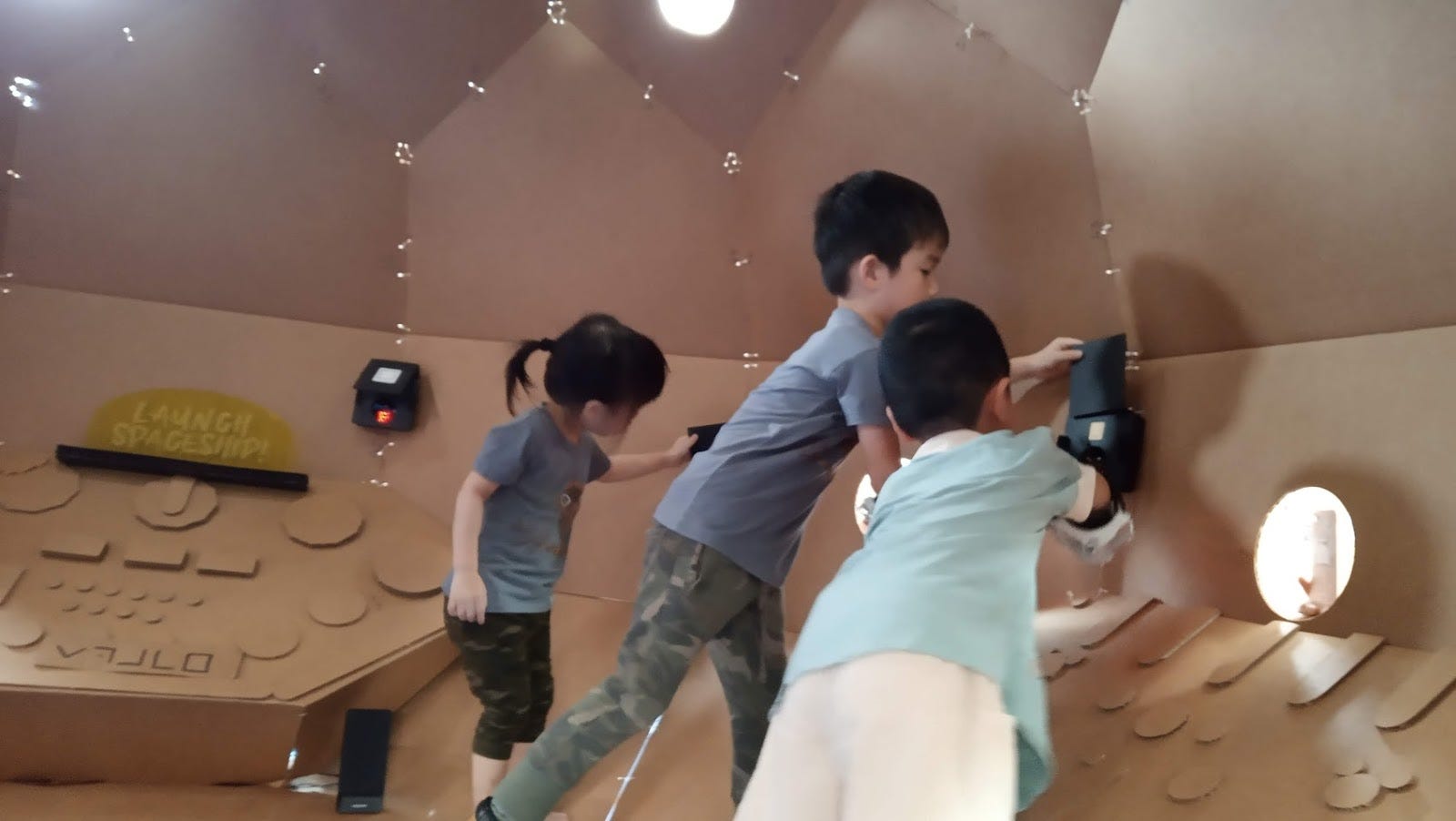 Children activate wall-mounted targets inside the cardboard spaceship game.