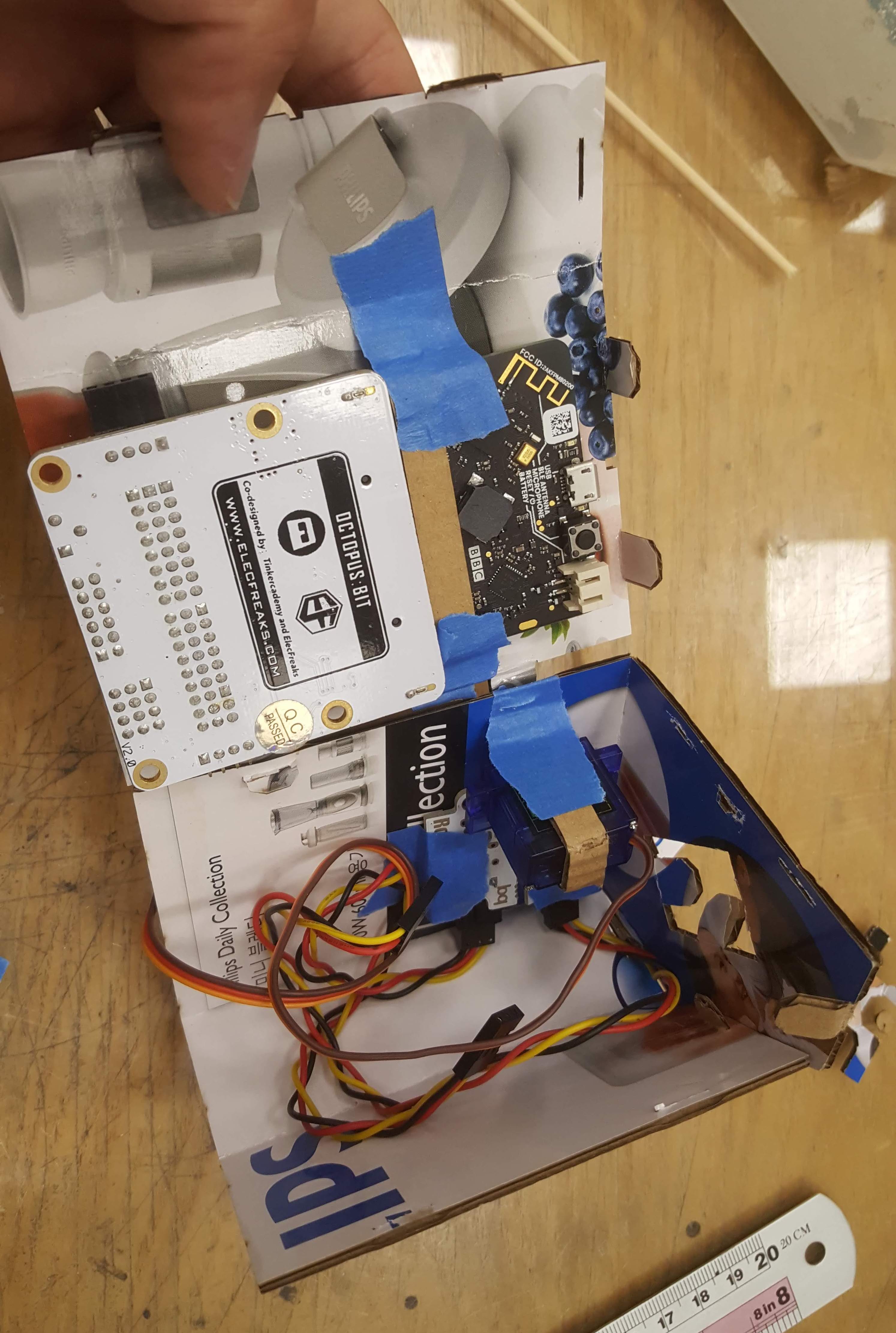 Hand-built cardboard electronics prototype assembled with folded tabs, cut slots, blue tape and exposed wiring.