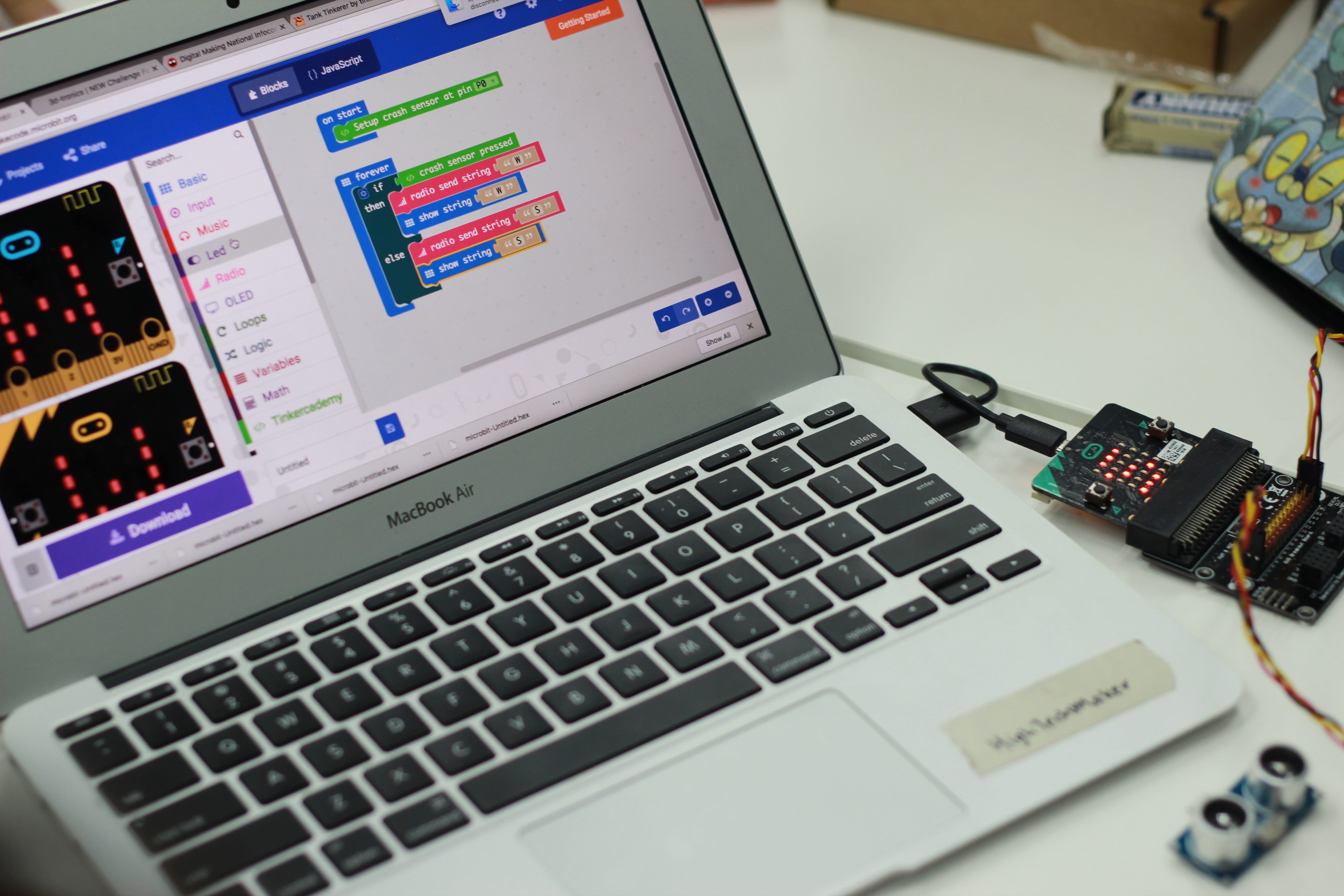Wired micro:bits displaying red LED patterns on a laptop running MakeCode blocks for the tank game.